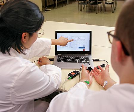 A photo taken from over the shoulders of two students. The students are wearing white labcoats and seated at a table in a lab environment. The student on the left is pointing at data on the screen of a laptop on the table. The student on the right has their forearms lying upturned on the table and they are connected to a Biopotential Sensor and a Finger Pulse Sensor.